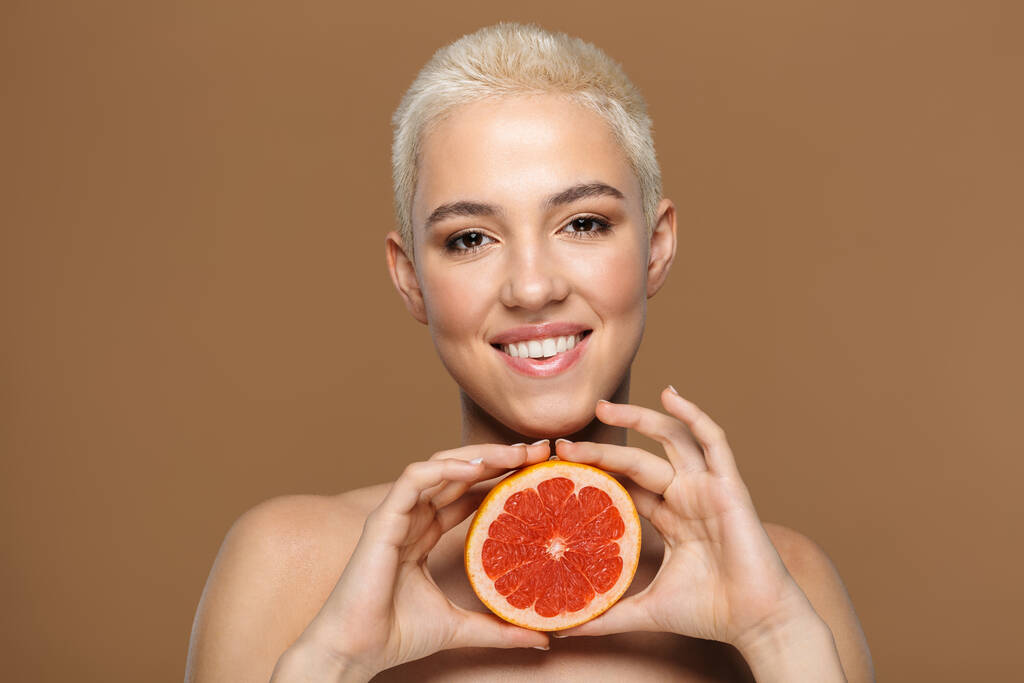 Afbeelding van een vrij vrolijke glimlachende jonge blonde stijlvolle vrouw met kort kapsel poseren geïsoleerd over donker beige muur achtergrond holding grapefruit. - Foto, afbeelding
