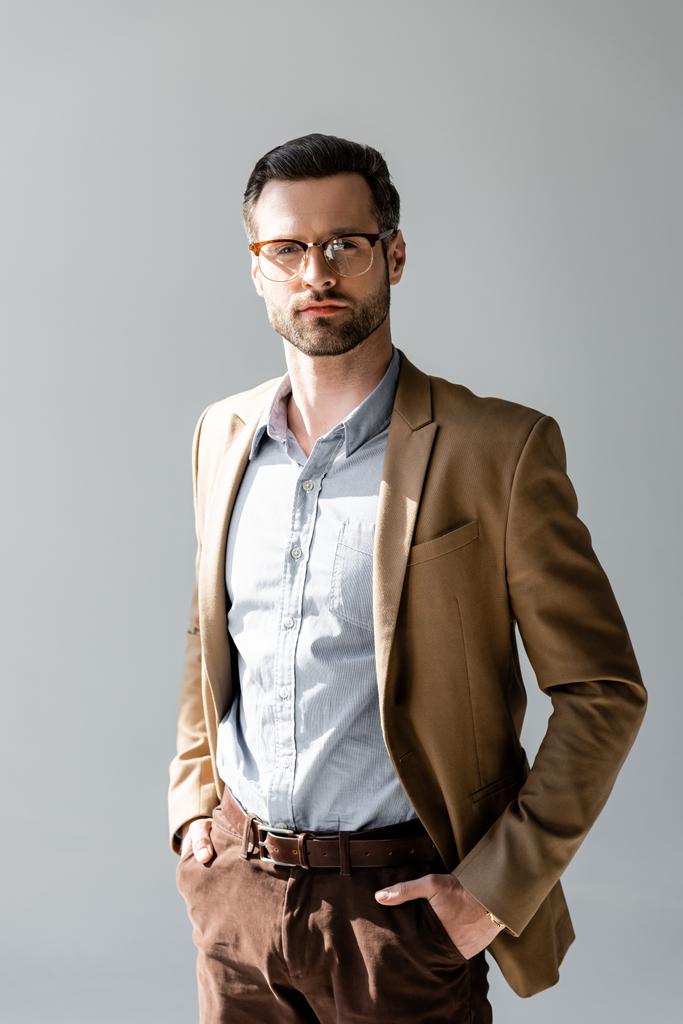 hombre de negocios guapo en gafas mirando a la cámara y de pie con las manos en bolsillos aislados en gris
  - Foto, imagen