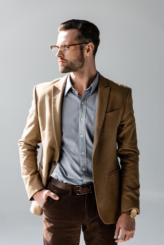 homem de negócios bonito em óculos olhando para longe e de pé com a mão no bolso isolado em cinza
  - Foto, Imagem