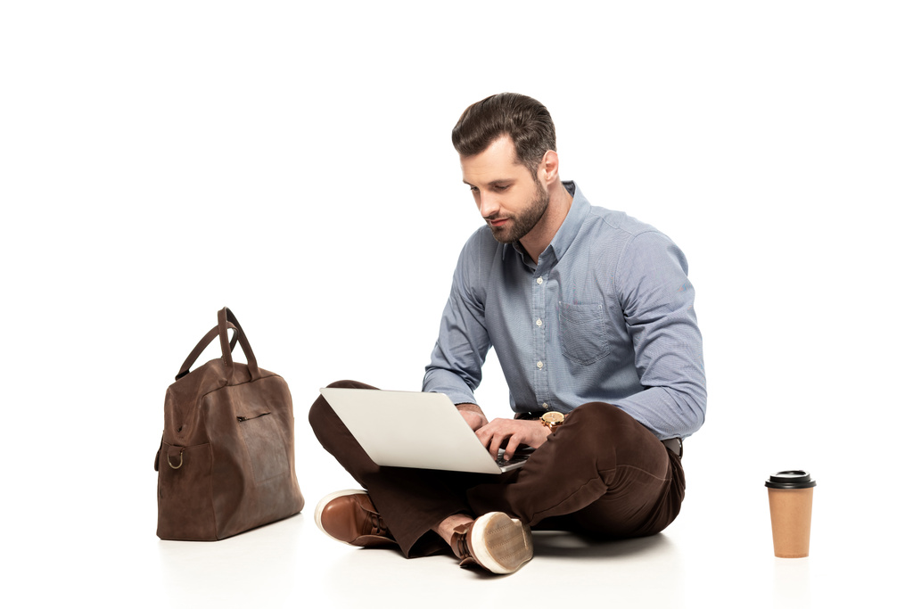 バッグや紙コップのそばに座りながらノートパソコンを使うハンサムなフリーランサー  - 写真・画像
