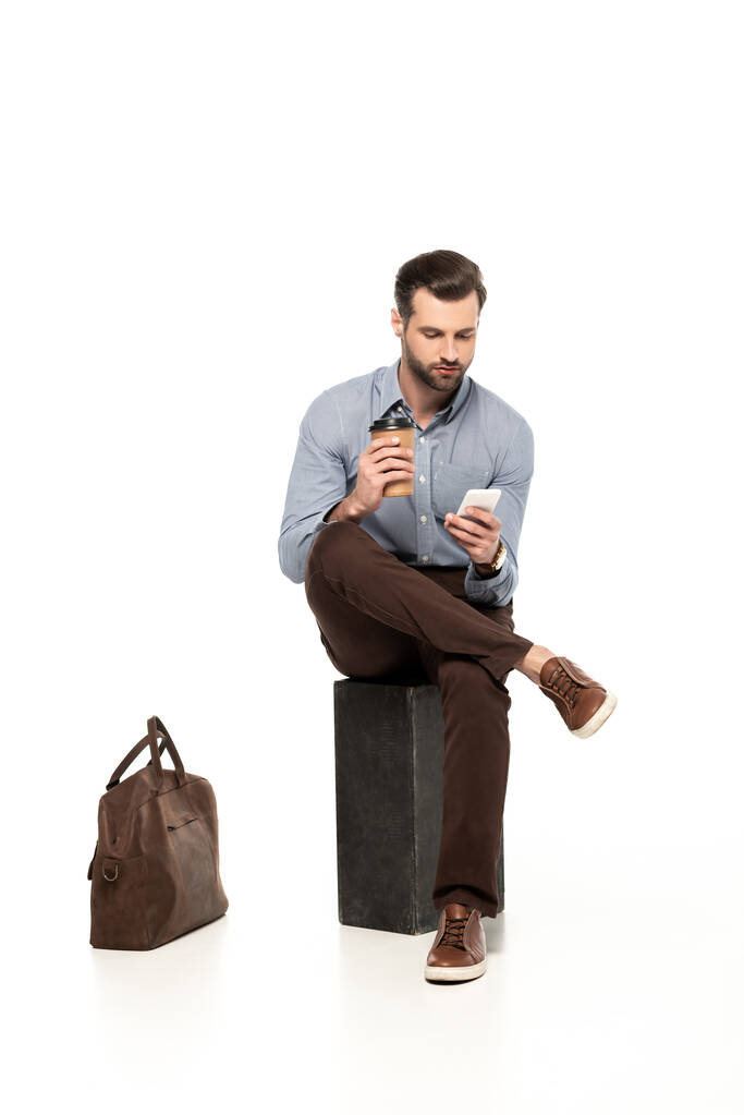 hombre de negocios guapo usando teléfono inteligente mientras está sentado cerca de la bolsa en blanco
  - Foto, imagen