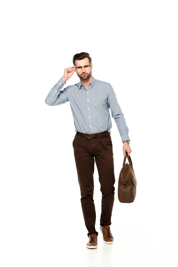 красивый и бородатый бизнесмен трогает очки и держит сумку, изолированную на белом
   - Фото, изображение