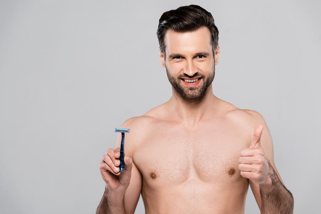handsome man holding razor while smiling and showing thumb up isolated on grey  - Photo, Image