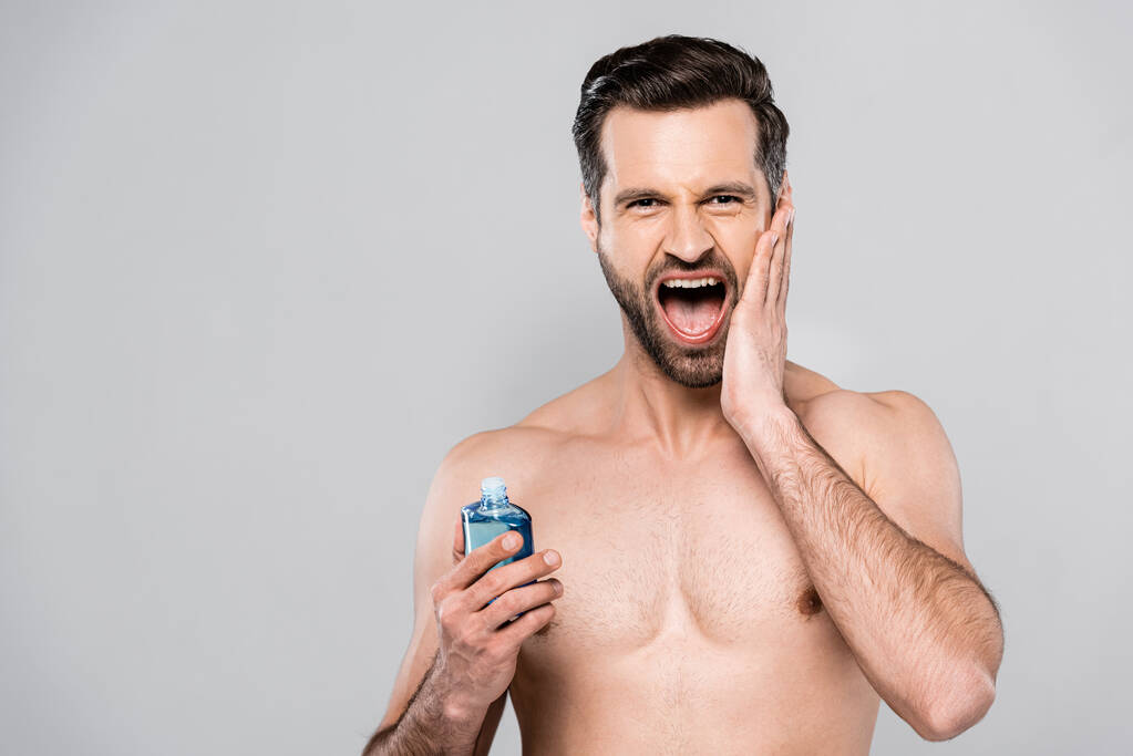 muskulöser Mann schreit, während er eine Flasche mit After Shave Lotion hält, isoliert auf grau  - Foto, Bild