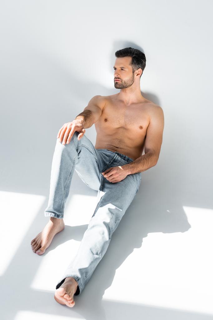 słońce na muskularnym człowieku w niebieskich dżinsach siedzi na szarości  - Zdjęcie, obraz