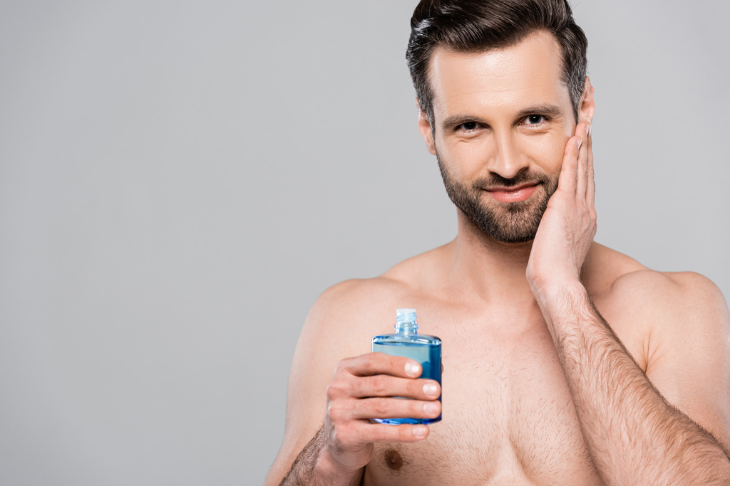happy man holding bottle with blue after shave lotion and touching face isolated on grey   - Photo, Image