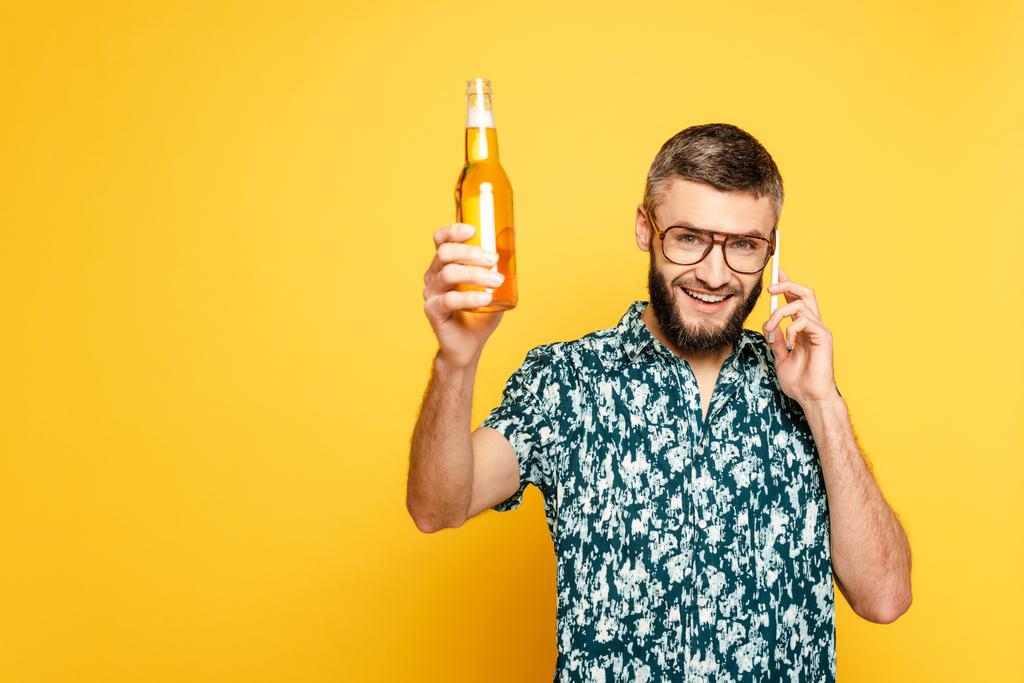 χαρούμενος γενειοφόρος τύπος σε ποτήρια με δροσιστικό μπουκάλι μπύρας μιλώντας στο smartphone σε κίτρινο - Φωτογραφία, εικόνα