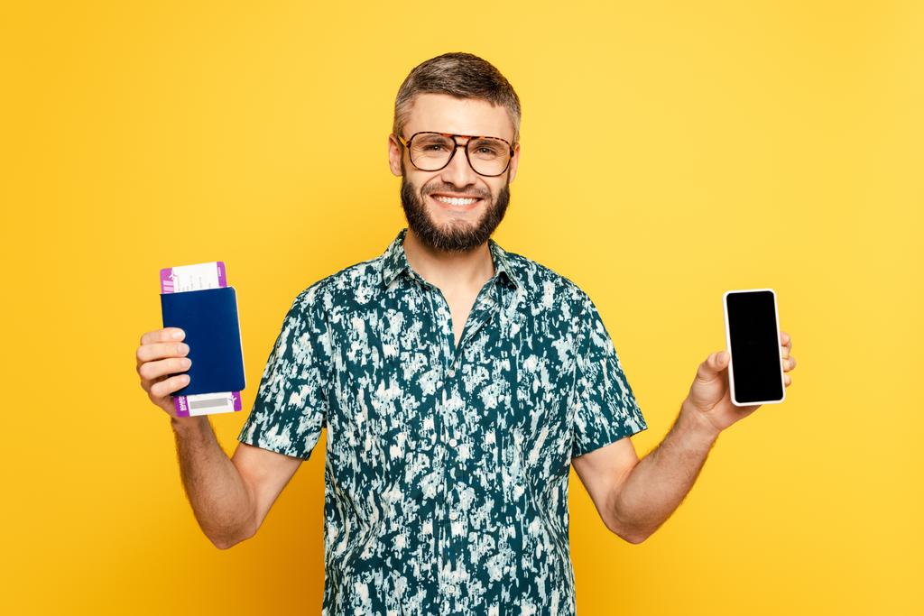 cara barbudo sorridente em óculos com passaporte e bilhete mostrando smartphone com tela em branco no amarelo
 - Foto, Imagem