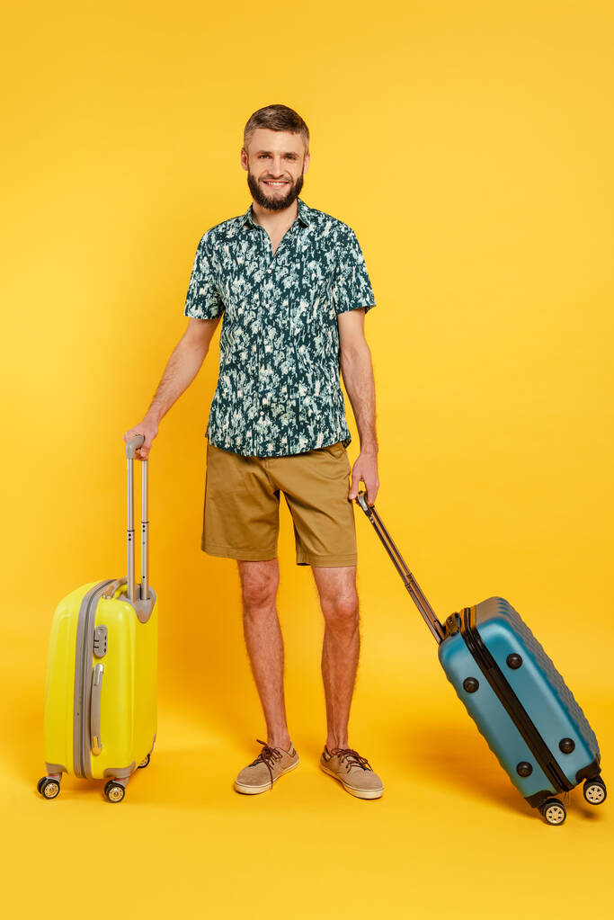 full length άποψη του ευτυχισμένος γενειοφόρος τύπος με ταξιδιωτικές τσάντες σε κίτρινο - Φωτογραφία, εικόνα