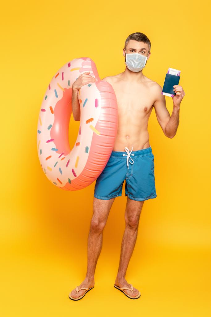 Full length άποψη του άντρα με μαγιό και ιατρική μάσκα με δαχτυλίδι κολύμπι και διαβατήριο σε κίτρινο - Φωτογραφία, εικόνα