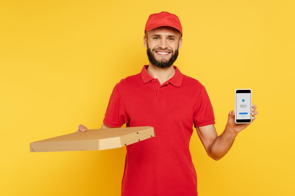 KYIV, UCRANIA - 30 de marzo de 2020: repartidor barbudo sonriente en uniforme rojo con caja de pizza que muestra el teléfono inteligente con aplicación de mensajero en amarillo
 - Foto, Imagen