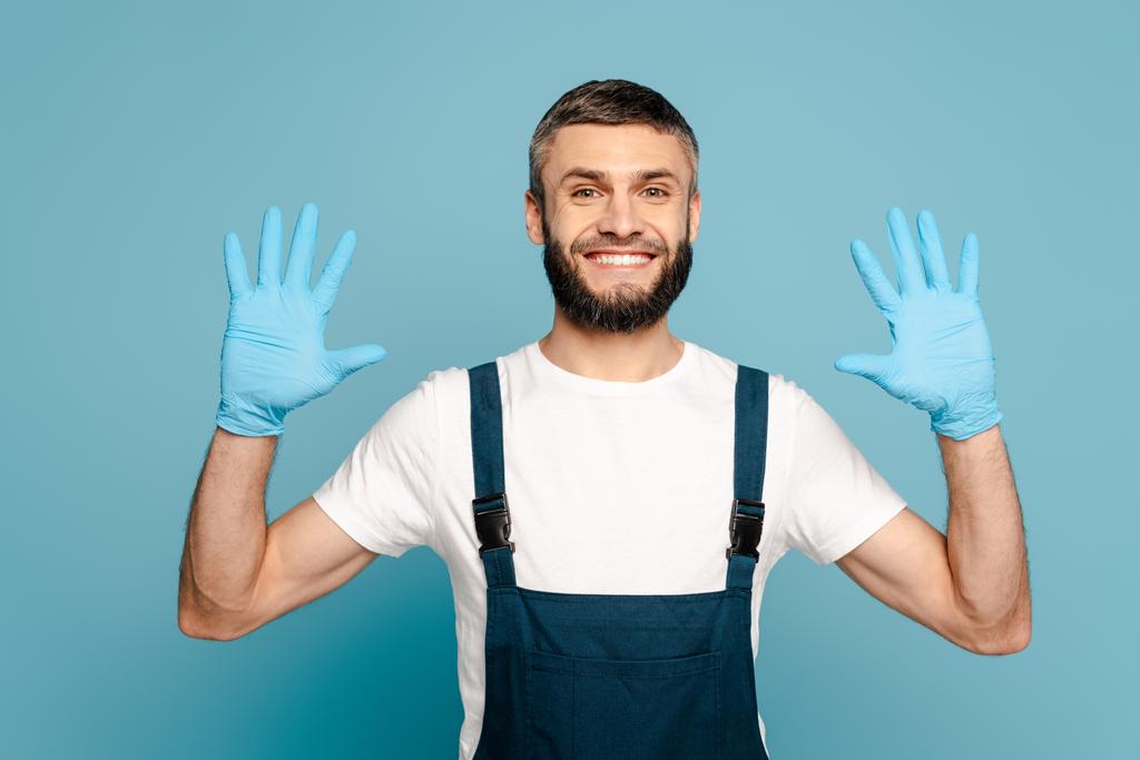 Mavi arka planda lastik eldivenli elleri gösteren üniformalı mutlu bir temizlikçi. - Fotoğraf, Görsel