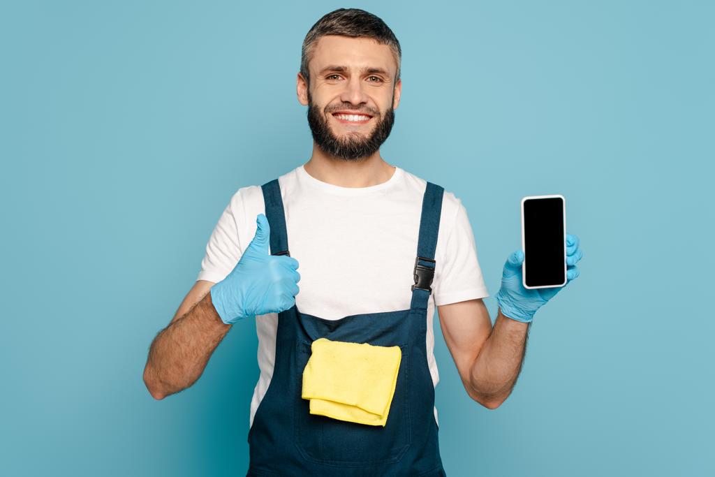 happy cleaner in uniform and rubber gloves with rug holding smartphone and showing thumb up on blue background - Photo, Image