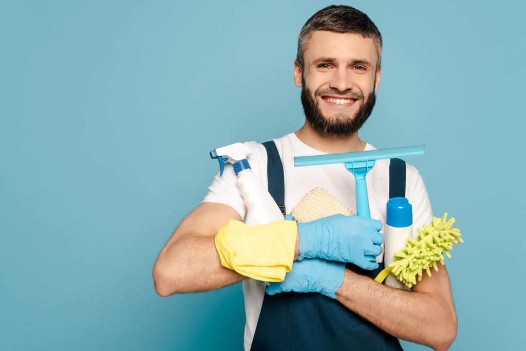 limpiador feliz en uniforme y guantes de goma que sostienen los suministros de limpieza sobre fondo azul
 - Foto, Imagen