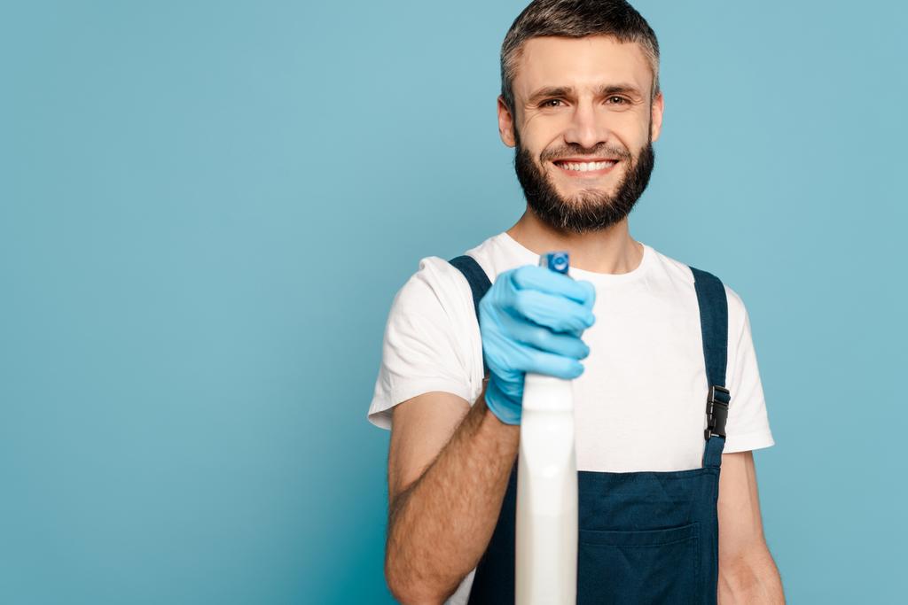fröhliche Reinigungskraft in Uniform und Gummihandschuhen mit Sprühwaschmittel auf blauem Hintergrund - Foto, Bild