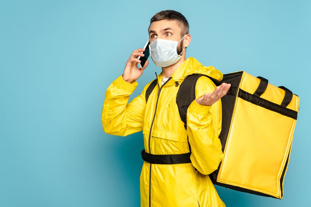 fattorino confuso in maschera medica e uniforme gialla con zaino che parla su smartphone su sfondo blu
 - Foto, immagini