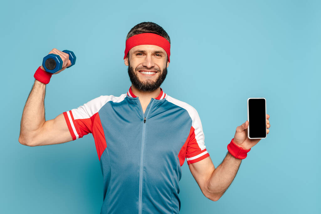 elegante sportivo sorridente che si esercita con manubrio e tenendo smartphone su sfondo blu
 - Foto, immagini