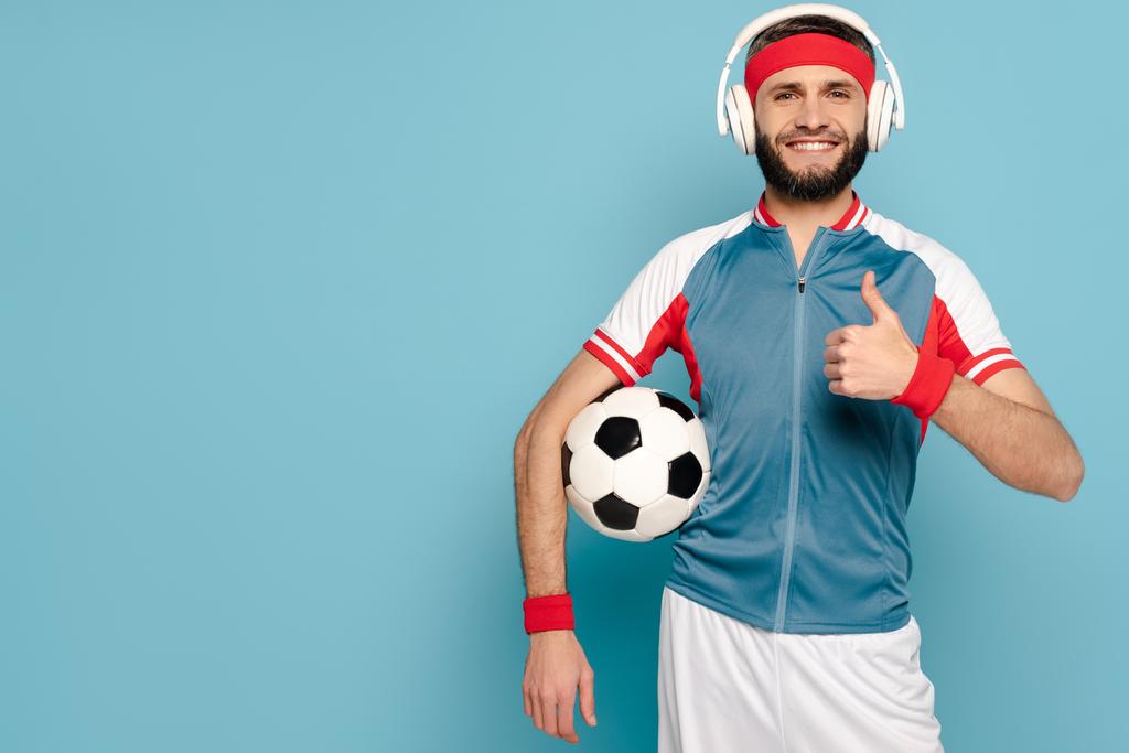 happy stylish sportsman in headphones with soccer ball showing thumb up on blue background - Photo, Image