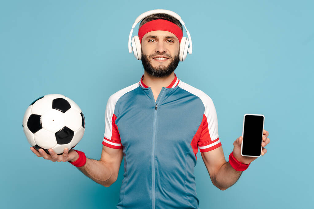 青い背景にサッカーボールとスマートフォンを持つヘッドフォンで幸せなスタイリッシュなスポーツマン - 写真・画像