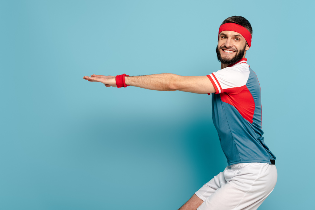 boční pohled na šťastný stylový sportovec dřepící na modrém pozadí - Fotografie, Obrázek