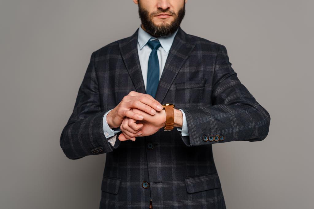 обрізаний вид бородатого бізнесмена в костюмі, який дивиться на наручний годинник на сірому фоні
 - Фото, зображення