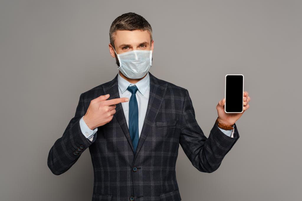 Tıbbi maskeli yakışıklı işadamı gri arka planda boş ekranlı akıllı telefonu işaret ediyor. - Fotoğraf, Görsel
