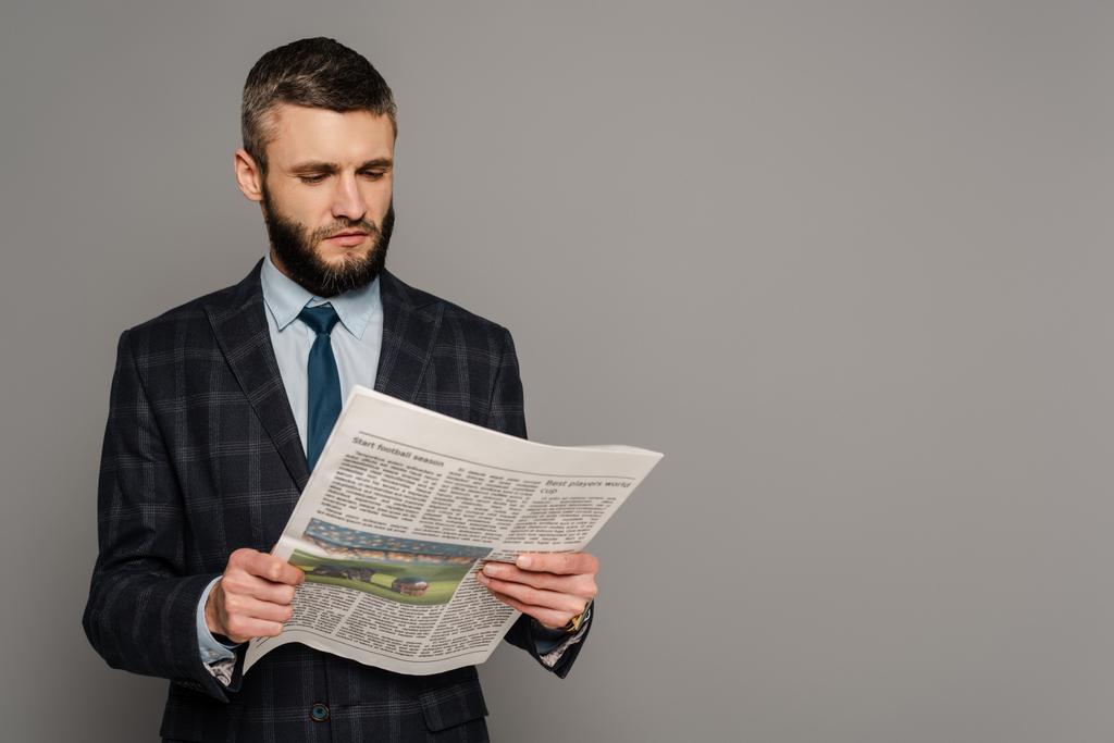 usmívající se pohledný vousatý podnikatel v obleku s novinami na šedém pozadí - Fotografie, Obrázek