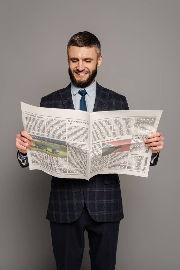 glimlachende knappe bebaarde zakenman in pak lezen krant op grijze achtergrond - Foto, afbeelding