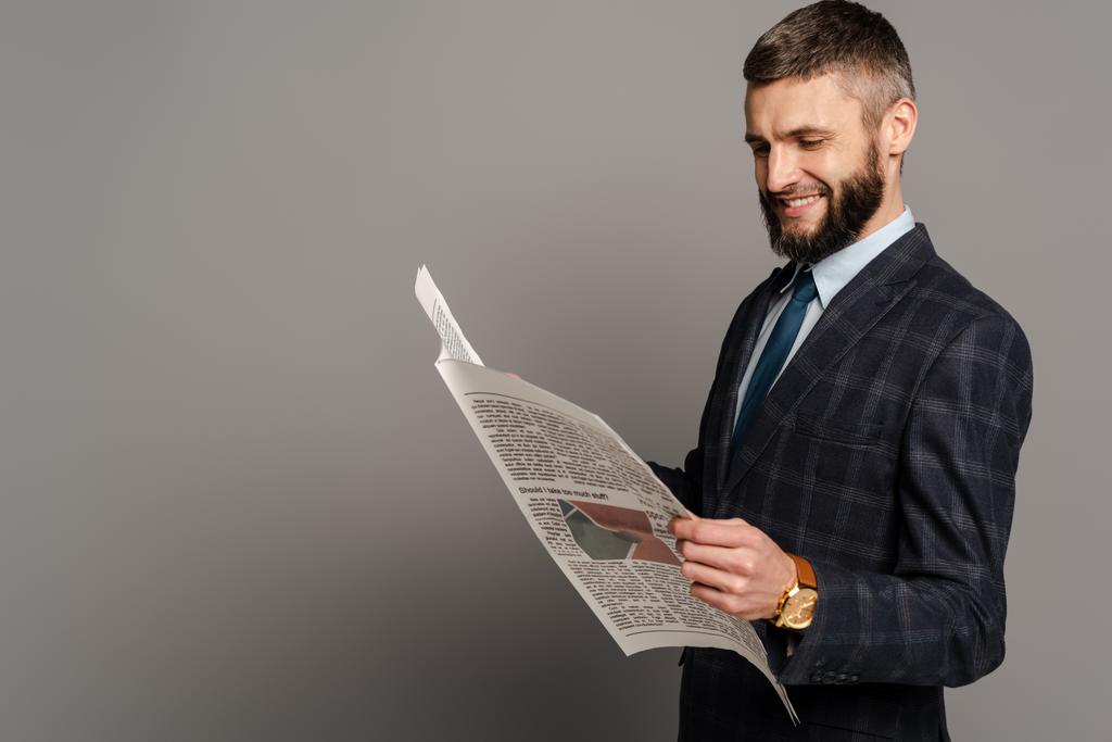 sonriente guapo barbudo hombre de negocios en traje leyendo periódico sobre fondo gris
 - Foto, imagen