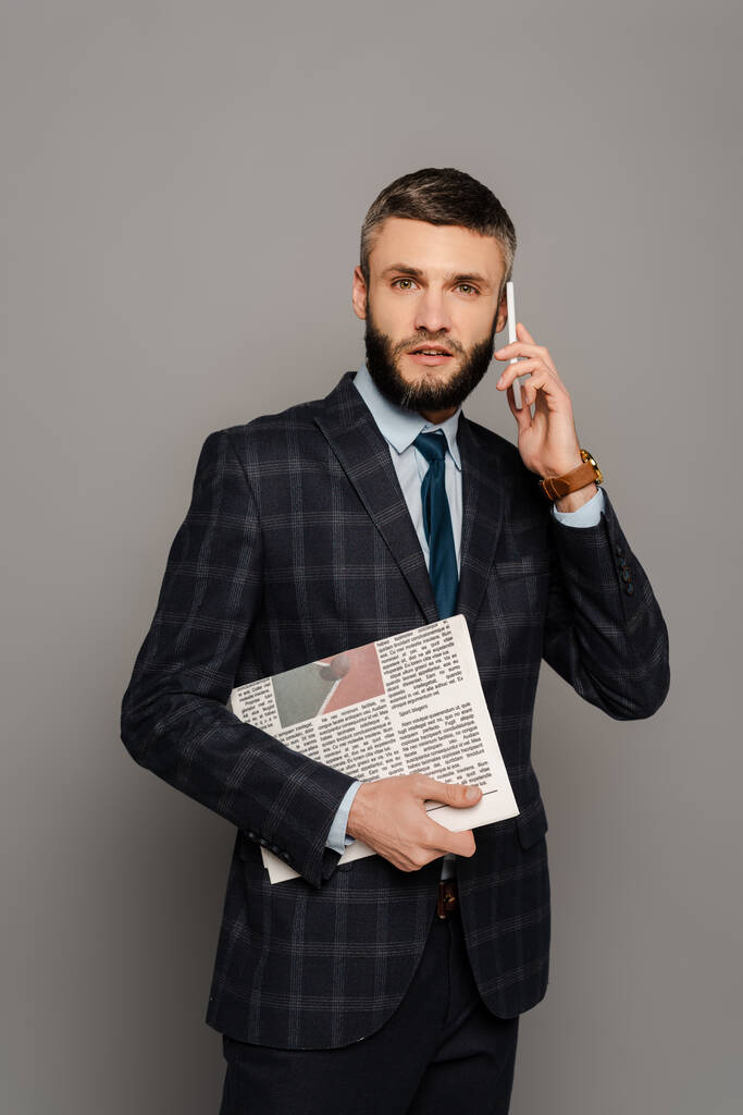 knappe bebaarde zakenman in pak met krant praten op smartphone op grijze achtergrond - Foto, afbeelding