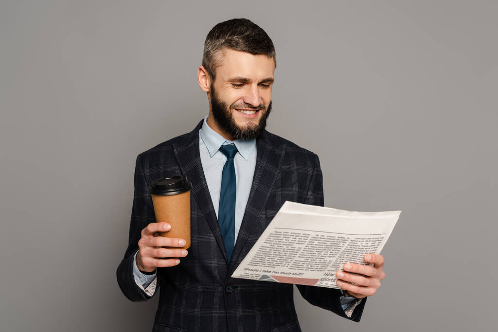 χαμογελαστός όμορφος γενειοφόρος επιχειρηματίας με κοστούμι και καφέ για να πάει να διαβάσει εφημερίδα σε γκρι φόντο - Φωτογραφία, εικόνα