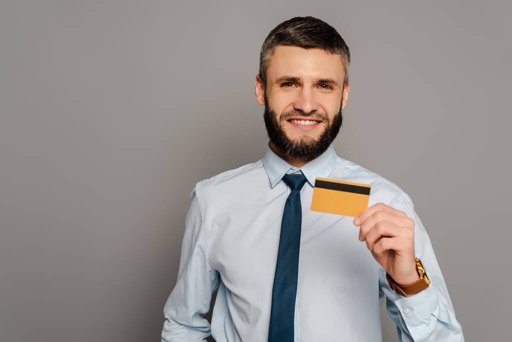 glimlachende knappe bebaarde zakenman met kredietkaart op grijze achtergrond - Foto, afbeelding