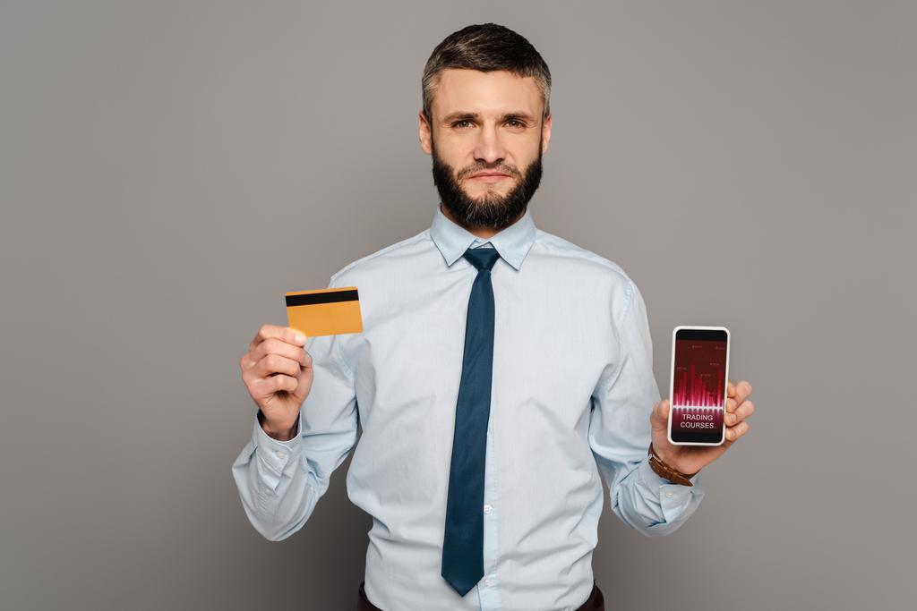 όμορφος γενειοφόρος επιχειρηματίας που κατέχει πιστωτική κάρτα και smartphone με εφαρμογή μαθημάτων συναλλαγών σε γκρι φόντο - Φωτογραφία, εικόνα