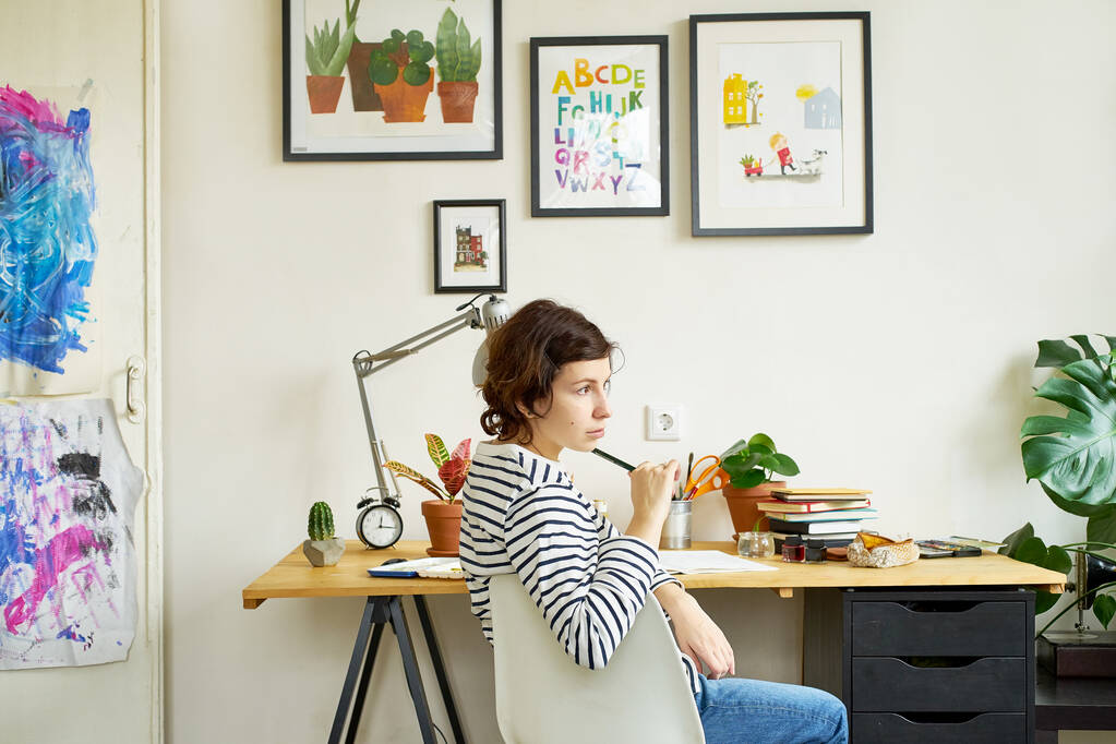 Artista feminina em seu local de trabalho trabalhando em casa. Jovem vestida de jeans e camisa listrada sentada à mesa e pensando. Procurando o conceito de inspiração
  - Foto, Imagem