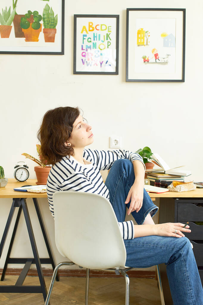 Vrouwelijke kunstenares op haar werkplek die thuis werkt. Vrouw gekleed in jeans en gestreept shirt zittend aan tafel en tekening illustratie. Op zoek naar inspiratie concept. - Foto, afbeelding