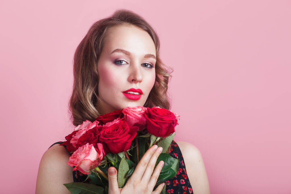 Piękna kobieta z kręconymi włosami i czerwonymi ustami trzymając bukiet róż na różowym tle studio - Zdjęcie, obraz
