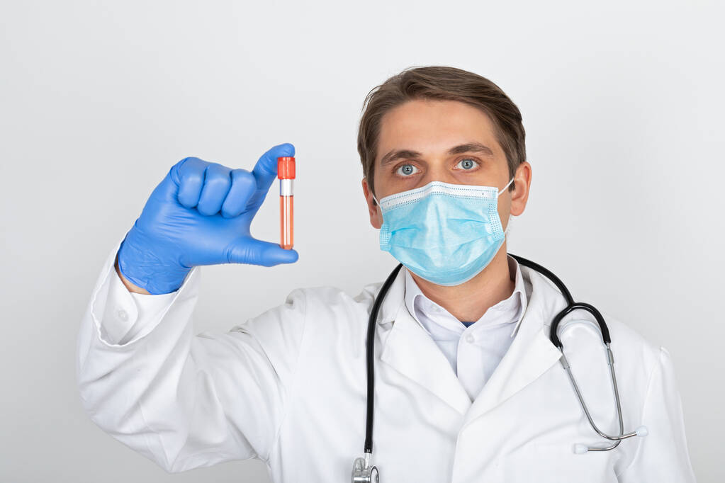 Cerrahi maskeli ve eldivenli genç bir erkek doktor beyaz arka planda kan örneği tutuyor. - Fotoğraf, Görsel