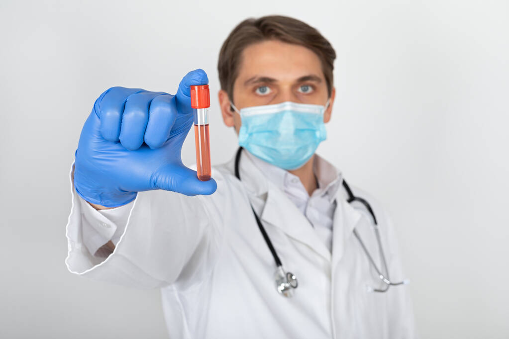 Cerrahi maskeli ve eldivenli genç bir erkek doktor beyaz arka planda kan örneği tutuyor. - Fotoğraf, Görsel