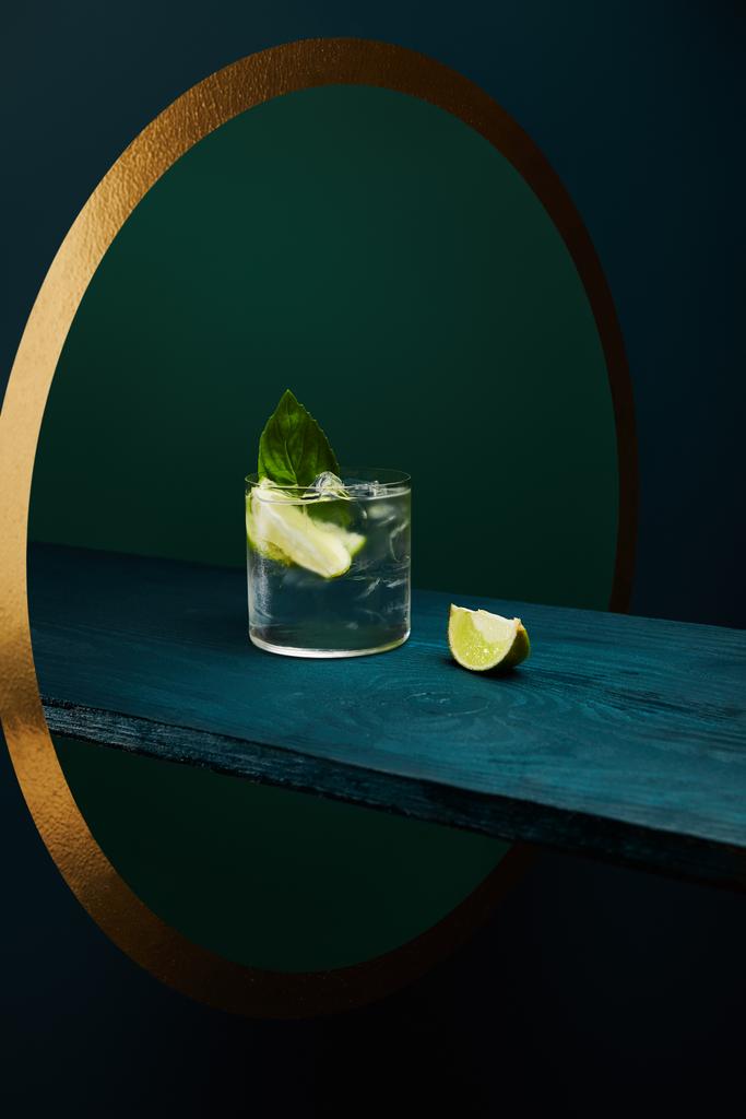 Bicchiere con bevanda rinfrescante con cubetti di ghiaccio, foglia di menta e fetta di lime su superficie di legno su sfondo geometrico blu e verde
 - Foto, immagini