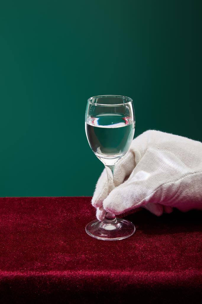 Ausgeschnittene Ansicht der Hand in Frotteehandschuh hält Schnapsglas mit Flüssigkeit isoliert auf grün - Foto, Bild