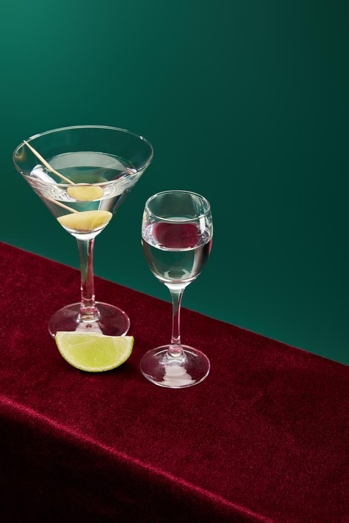 Hochwinkelaufnahme von Schnaps- und Cocktailglas mit Wermut, Limettenscheibe und ganzen Oliven auf Zahnstocher auf roter Velouroberfläche isoliert auf grünem Grund - Foto, Bild