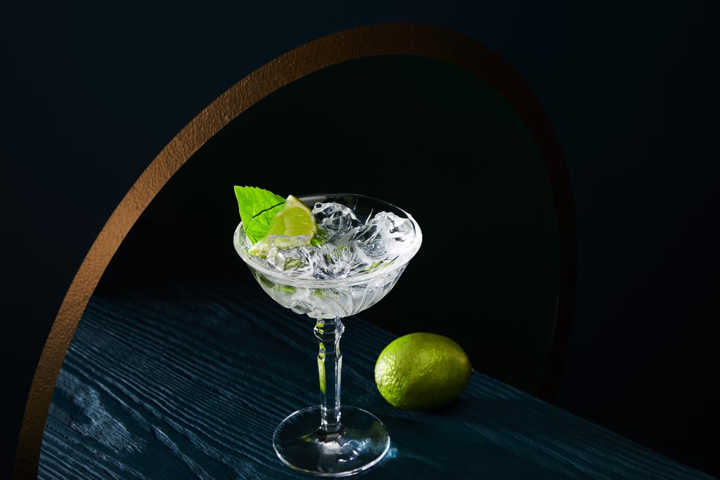 high angle view of cocktail glass with ice, mint leaf and whole lime on blue wooden surface on geometric background with golden circle - Photo, Image