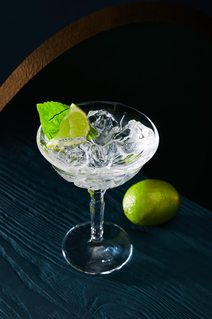 high angle view of cocktail glass with ice, mint leaf and whole lime on blue wooden background - Photo, Image