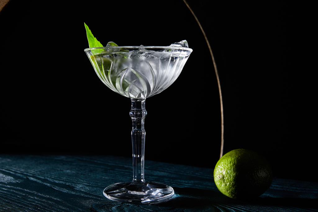 verre à cocktail avec boisson avec glaçons, feuille de menthe et citron vert entier sur une surface en bois bleu sur fond noir avec des lignes géométriques
 - Photo, image