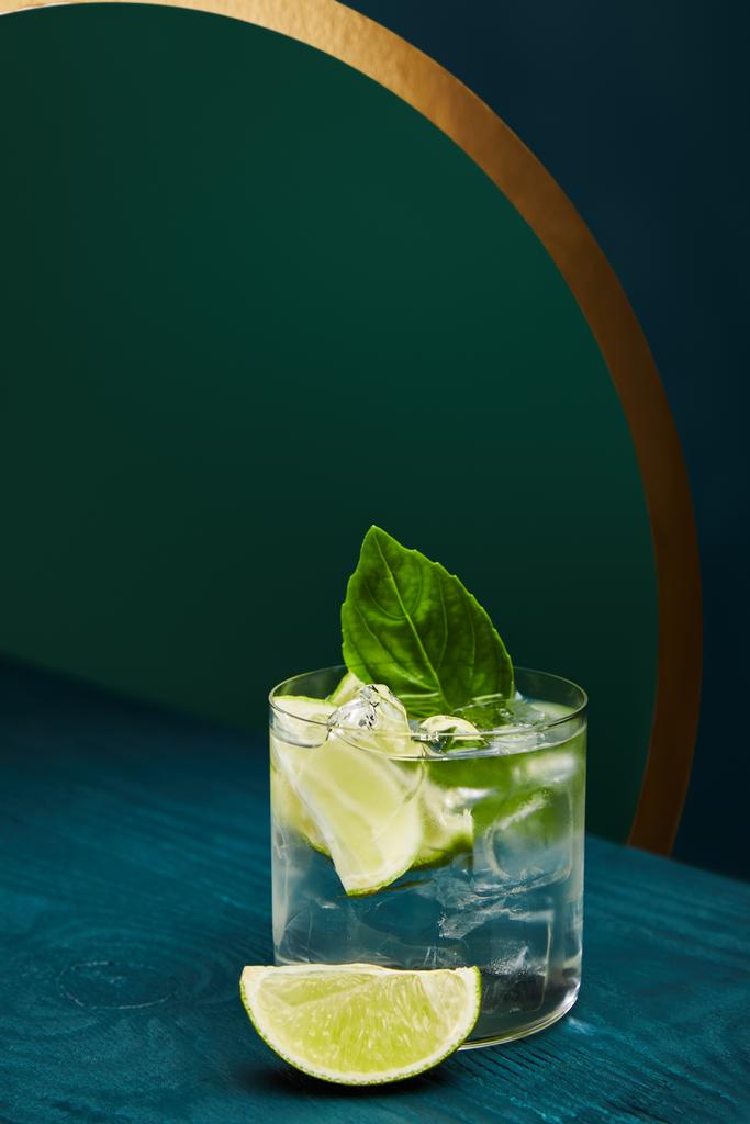 old fashioned glass with fresh drink, mint leaf and lime slice on blue wooden surface on green and blue geometric background with golden lines - Photo, Image