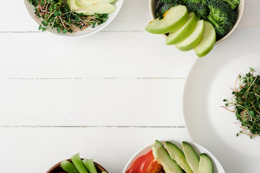 вид сверху на свежие овощи и фрукты с микрозеленым в мисках на белой деревянной поверхности
 - Фото, изображение