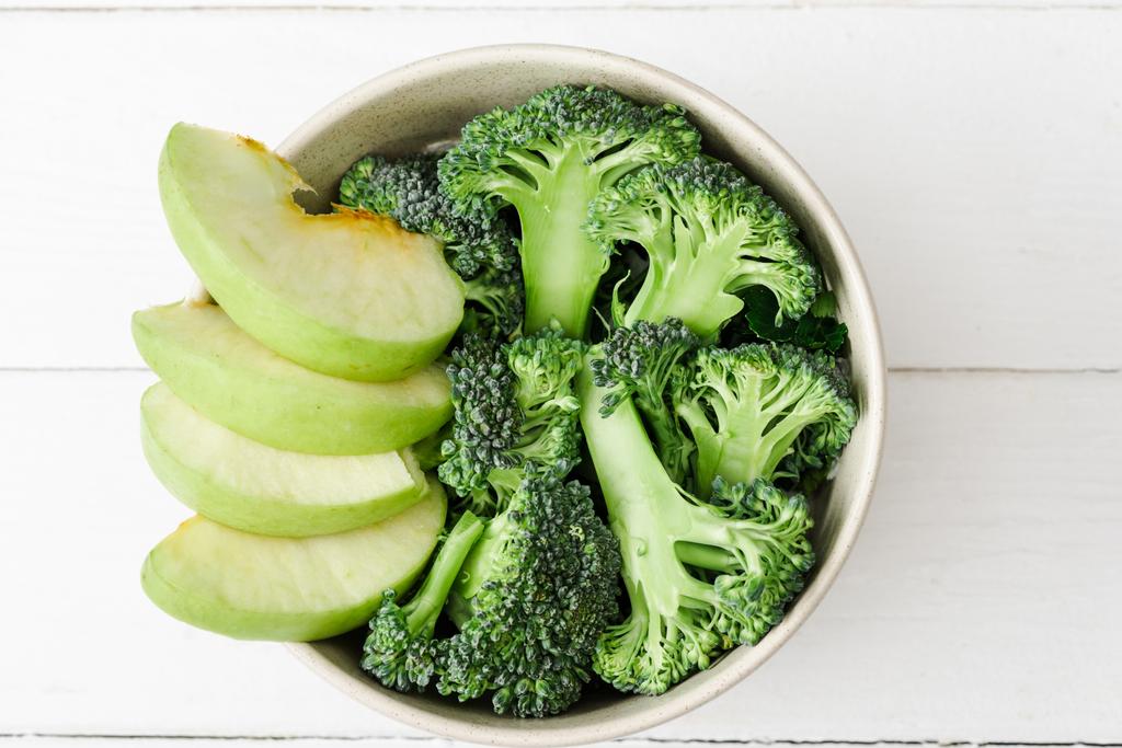 vrchní pohled na čerstvé zelené jablko a brokolici v misce na bílém dřevěném povrchu - Fotografie, Obrázek