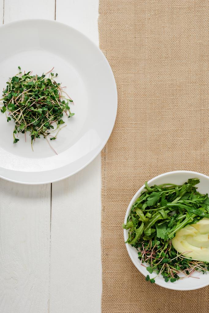 Draufsicht auf frisches Mikrogrün auf Teller neben Schüssel mit grünem Salat auf beiger Serviette auf weißer Holzoberfläche - Foto, Bild