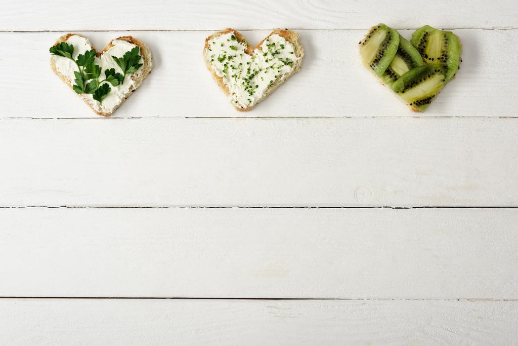 κορυφαία άποψη του canape σε σχήμα καρδιάς με κρεμώδες τυρί, μικροπράσινο, μαϊντανό και ακτινίδιο σε λευκή ξύλινη επιφάνεια - Φωτογραφία, εικόνα