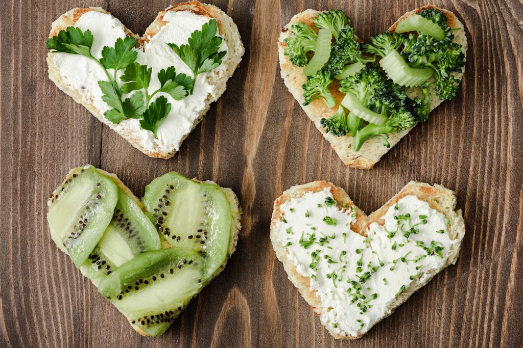 top view of heart shaped canape with creamy cheese, broccoli, microgreen, parsley and kiwi on wooden surface - Photo, Image